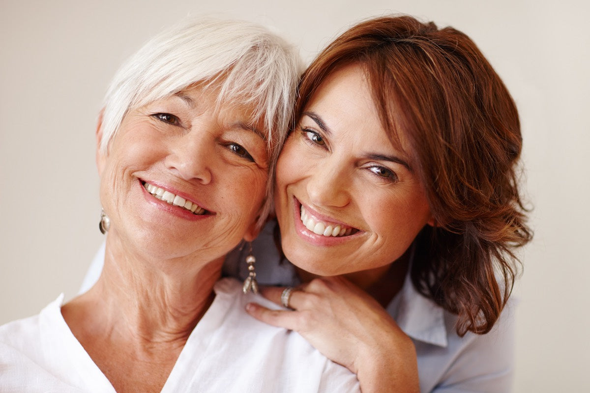 A white-haired woman smiles at the camera while a dark-haired woman hovers lovingly behind her, also smiling